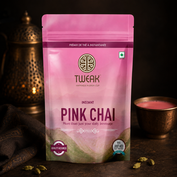 A stand-up pouch of Tweak Instant Pink Chai tea, featuring a pink and magenta gradient design with the brand logo and product name. The pouch is displayed on a dark wooden surface with a brass teapot in the background and a small cup of pink tea on the right.